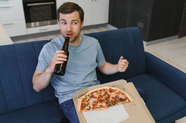 TV şovu izlerken pizza yiyor. İçeride sakallı bir adam.