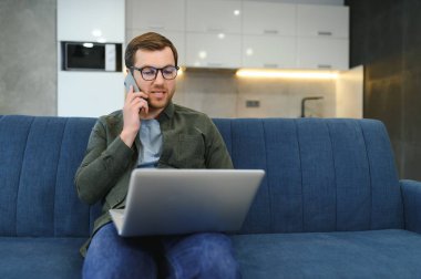 Neşeli, meşgul, genç, beyaz erkek yönetici kanepede bilgisayarla oturur, telefonla konuşur, müşteriyle konuşur, beyaz oda içlerindeki boş alana bakar. İş, evde uzaktan çalışma, reklam ve teklif