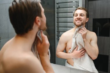 Young Caucasian man look in mirror do skincare facial routine after shower in home bathroom, millennial male in towel after bath apply face lotion or cream on skin after shaving, hygiene concept