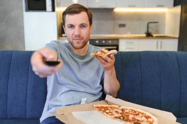 TV şovu izlerken pizza yiyor. İçeride sakallı bir adam.