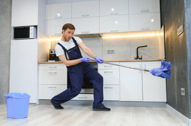 Profesyonel bir temizlikçi olarak mavi üniformalı bir adam dairenin oturma odasında paspas çubuğu ve kovayla yerleri yıkıyor..