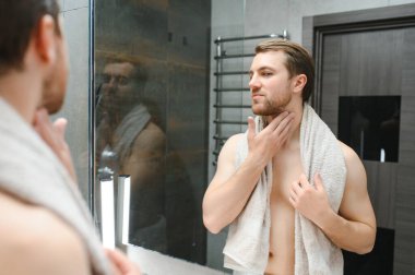 Young Caucasian man look in mirror do skincare facial routine after shower in home bathroom, millennial male in towel after bath apply face lotion or cream on skin after shaving, hygiene concept