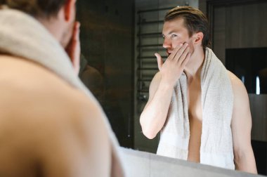 Young Caucasian man look in mirror do skincare facial routine after shower in home bathroom, millennial male in towel after bath apply face lotion or cream on skin after shaving, hygiene concept