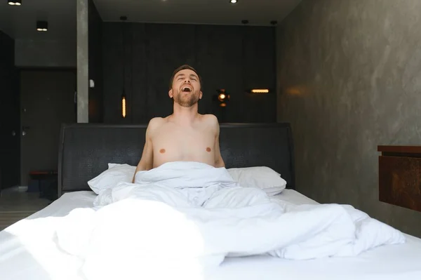 stock image Young man waking up and rise hands stretching on white bed in the morning with sunlight through window.