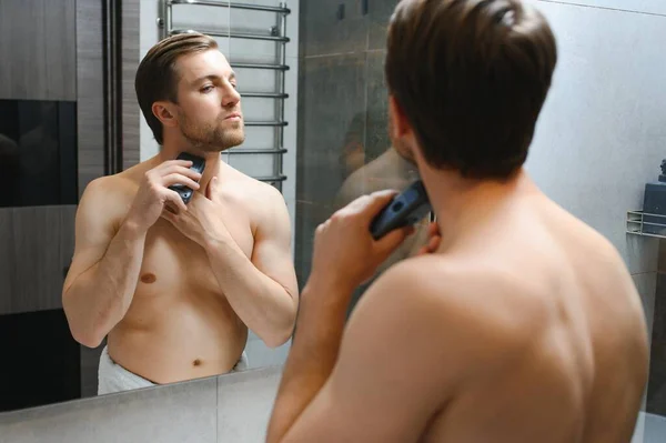 Reflection of young man in mirror shaving with electric shaver.