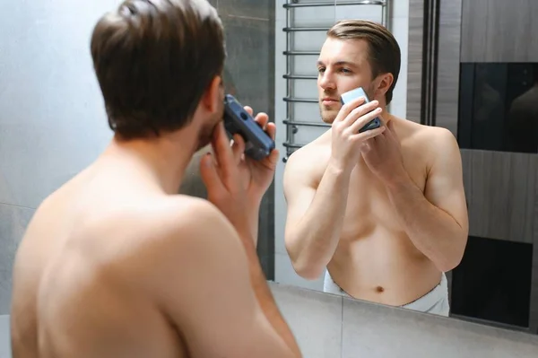 仪容仪表和人们的观念 关闭的年轻人期待着镜子刮胡子与微调或电动剃须刀在家里浴室 — 图库照片