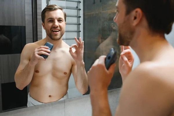 Reflection of young man in mirror shaving with electric shaver.