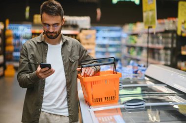 Süpermarkette: yakışıklı adam akıllı telefon kullanan ve beslenme bakar konserveler değerini. O ile alışveriş sepeti konserve ürünleri bölümünde duruyor