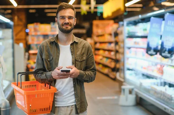 At the Supermarket: Handsome Man Uses Smartphone and Looks at Nutritional Value of the Canned Goods. He\'s Standing with Shopping Cart in Canned Goods Section