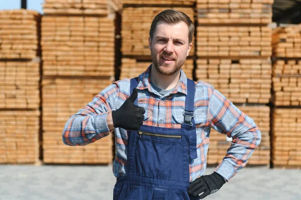 Portrait of a handsome worker choosing the best wooden boards. Carpenter standing next to a big stack of wood bars in a warehouse
