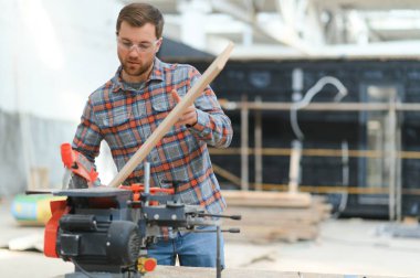 Profesyonel marangoz kereste fabrikasında tahta kesmek için testere makinesi kullanıyor. Marangozluk atölyesinde elektrikli testere ile çalışan yetenekli bir dolap yapımcısı. Erkek katılımcı, ahşap üretim tezgahı