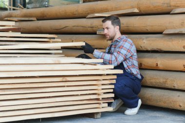 İnşaatçının elinde tahta kalaslar var. Konsept - kereste satışı. İnşaatçı ahşap tahtaları düşünüyor. Tahta işleme işi. Tahta kereste yığını.