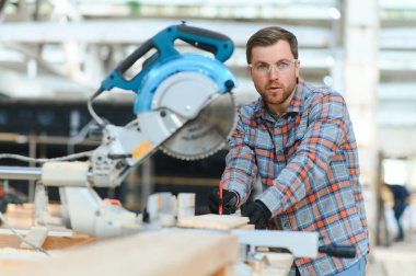 Bir marangoz, makine aletinin ahşap işçiliğinde çalışır. Adam mobilya kutuları topluyor. Mobilya detaylarını dairesel testere ile kesiyor. Parça parça kesme işlemi. Atölyenin arka planına karşı