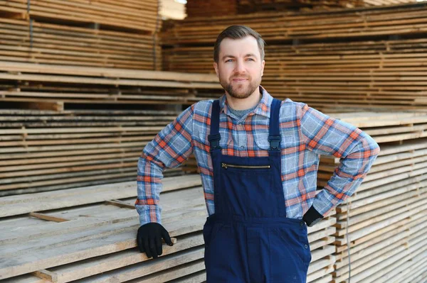 stock image Portrait of a handsome worker choosing the best wooden boards. Carpenter standing next to a big stack of wood bars in a warehouse