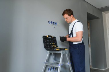 Elektrik İmalathanesi, prizler ve şalter kurulumu. Elektrikçi aletli profesyonel bir işçi. Tamir alanının arka planına karşı