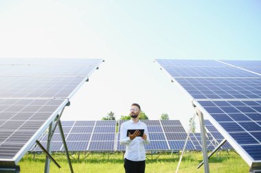 Beyaz gömlekli genç mühendis elinde bir tablet ve güneş enerjisi santralindeki fotovoltaik panelleri doğruluyor. Metnin için boşluk kopyala.