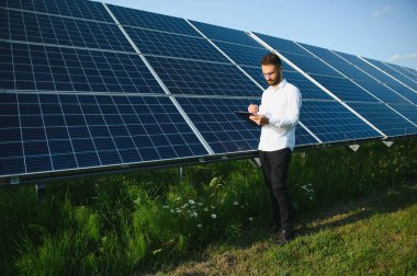 Kırsal kesimde güneşli bir günde beyaz gömlekli bir adam fotovoltaik panellerin yanında duruyor..