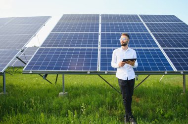 Güneşli bir günde güneş çiftliğinde çalışırken fotovoltaik panelleri kontrol etmek için tablet kullanan sakallı erkek mühendis..