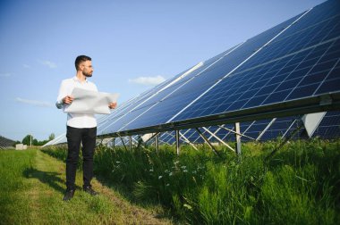 Kırsal kesimde güneşli bir günde beyaz gömlekli bir adam fotovoltaik panellerin yanında duruyor..
