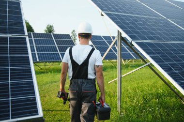Elinde matkap, elinde tornavida olan üniformalı bir erkek işçi güneş panellerinin yanındaki bir merdiven üzerinde..