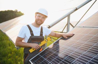 Erkek işçilerin güneş modülleri kurarken ve fotovoltaik güneş dizisini desteklerken yan görünümü. Güneş paneliyle çalışırken kaskını takan bir elektrikçi. Güneş enerjisi kavramı.