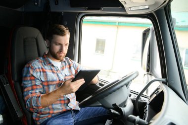 Kamyon şoförü arabanın direksiyonunda oturuyor ve elinde dijital tablet tutuyor..
