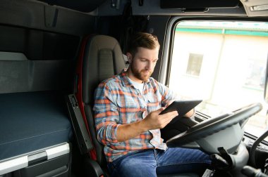 Kamyon şoförü arabanın direksiyonunda oturuyor ve elinde dijital tablet tutuyor..