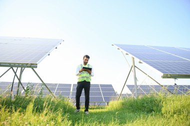 Güneş panellerinin yanında duran tabletli genç erkek mühendis, temiz mavi gökyüzü geçmişe sahip tarım arazisi, yenilenebilir enerji, temiz enerji