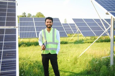 Genç Hintli teknisyen ya da yönetici güneş panelinin yanında duran resmi giysiler giyiyor. Yenilenebilir enerji, çapraz duran adam, boşluğu kopyala.