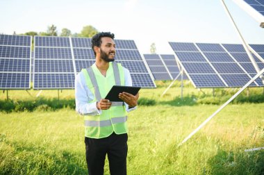 Genç Hintli teknisyen ya da yönetici güneş panelinin yanında duran resmi giysiler giyiyor. Yenilenebilir enerji, çapraz duran adam, boşluğu kopyala.