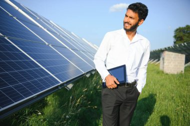 Güneş panellerinin yanında duran tabletli genç erkek mühendis, temiz mavi gökyüzü geçmişe sahip tarım arazisi, yenilenebilir enerji, temiz enerji