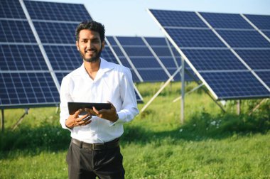 Genç Hintli erkek mühendisin portresi güneş panellerinin yanında duruyor, açık mavi gökyüzü arka planı, yenilenebilir ve temiz enerjisi var. beceri Hindistan, kopyalama alanı