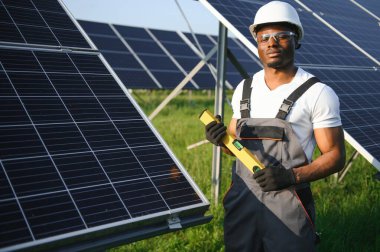 Beyaz kasklı ve gri tulumlu Afro-Amerikan adam güneş panellerinin arasında duruyor..