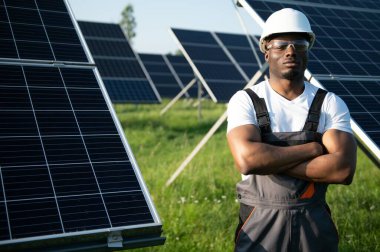 Beyaz kasklı ve gri tulumlu Afro-Amerikan adam güneş panellerinin arasında duruyor..