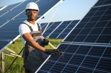 Güvenlik kaskı ve gözlüklü Afrikalı Amerikalı adam güneş panellerini tornavidayla sıkıyor. İstasyonda servis çalışması yaparken araçları kullanan yetkili teknisyen.