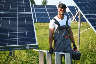 Beyaz kasklı ve gri tulumlu Afro-Amerikan adam güneş panellerinin arasında duruyor..