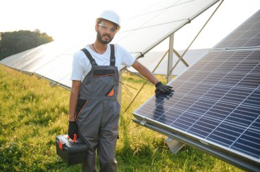 Kasklı ve kahverengi tulumlu erkek Arap mühendis güneş panellerindeki direnci kontrol ediyor. İstasyonda çalışan Hintli adam.