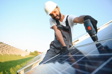 Kasklı ve kahverengi tulumlu erkek Arap mühendis güneş panellerindeki direnci kontrol ediyor. İstasyonda çalışan Hintli adam.