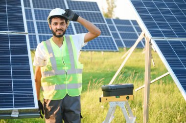 Kasklı ve kahverengi tulumlu erkek Arap mühendis güneş panellerindeki direnci kontrol ediyor. İstasyonda çalışan Hintli adam.