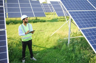 Genç Hintli teknisyen ya da yönetici güneş panelinin yanında duran resmi giysiler giyiyor. Yenilenebilir enerji, çapraz duran adam, boşluğu kopyala.