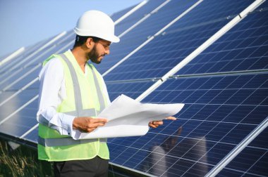 Mavi gökyüzüne karşı güneş panelleri yakınında duran beyaz başlıklı genç Hintli teknisyen portresi. Endüstriyel işçi güneş sistemi kurulumu, yenilenebilir yeşil enerji üretimi konsepti.