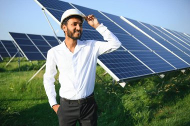 Mavi gökyüzüne karşı güneş panelleri yakınında duran beyaz başlıklı genç Hintli teknisyen portresi. Endüstriyel işçi güneş sistemi kurulumu, yenilenebilir yeşil enerji üretimi konsepti.