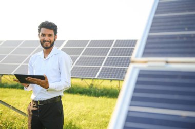 Genç Hintli teknisyen ya da yönetici güneş panelinin yanında duran resmi giysiler giyiyor. Yenilenebilir enerji, çapraz duran adam, boşluğu kopyala.
