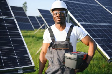 Gri tulumlu ve turuncu başlıklı ehil enerji mühendisi sahada yürürken güneş panellerini kontrol ediyor. Elinde enstrümanlarla pano ve konteynır taşıyan Afrikalı Amerikalı bir adam..