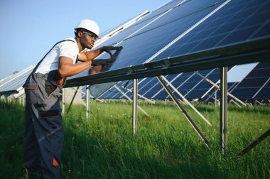 Beyaz kasklı ve gri tulumlu Afro-Amerikan adam güneş panellerinin arasında duruyor..