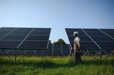 Beyaz kasklı ve gri tulumlu Afro-Amerikan adam güneş panellerinin arasında duruyor..