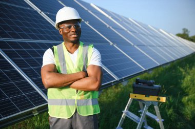 Gri tulumlu ve turuncu başlıklı ehil enerji mühendisi sahada yürürken güneş panellerini kontrol ediyor. Elinde enstrümanlarla pano ve konteynır taşıyan Afrikalı Amerikalı bir adam..