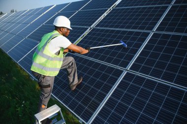 Üniformalı alternatif enerji santrali işçisi güneş panellerini paspasla temizliyor. Yakışıklı Afro-Amerikalı ekipmanlarla ilgileniyor.