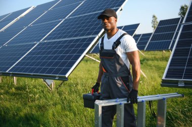 Gri tulumlu ehil enerji mühendisi ve beyaz miğfer sahada yürürken güneş panellerini kontrol ediyor. Elinde enstrümanlarla pano ve konteynır taşıyan Afrikalı Amerikalı bir adam..