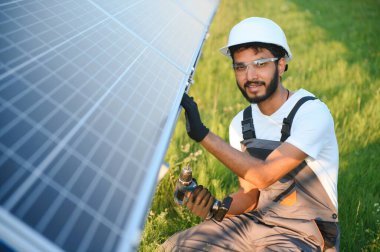 Kasklı ve kahverengi tulumlu erkek Arap mühendis güneş panellerindeki direnci kontrol ediyor. İstasyonda çalışan Hintli adam.